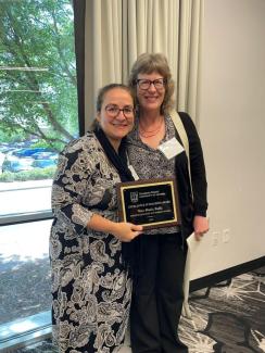 Photo of two women standing in front of a large window. The woman on the left is holding a plaque commemorating her Excellence in Teaching Award